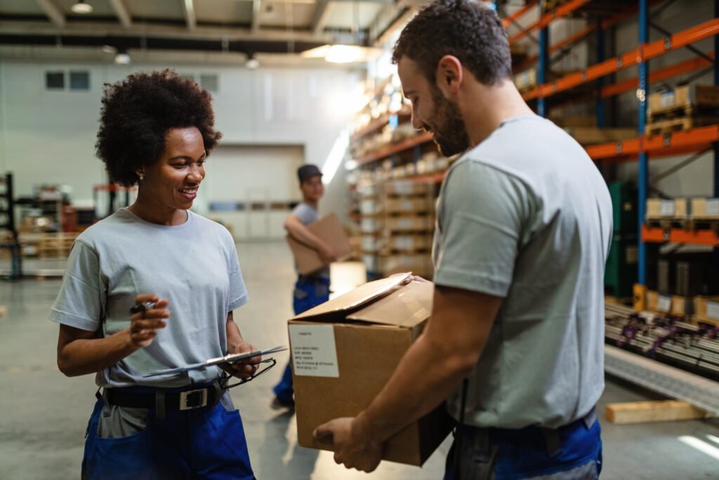 happy-black-female-worker-going-through-check-list-and-talking-to-her-coworker-who-is-carrying-cardboard-box-in-distribution-warehouse